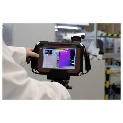 A person in a lab coat holds a thermal imaging device displaying temperature variations on a screen in an industrial setting.