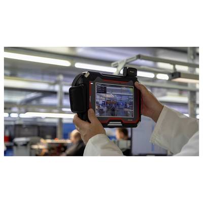 Person in lab coat holds a tablet device displaying a digital interface, capturing a live image of a manufacturing facility interior.