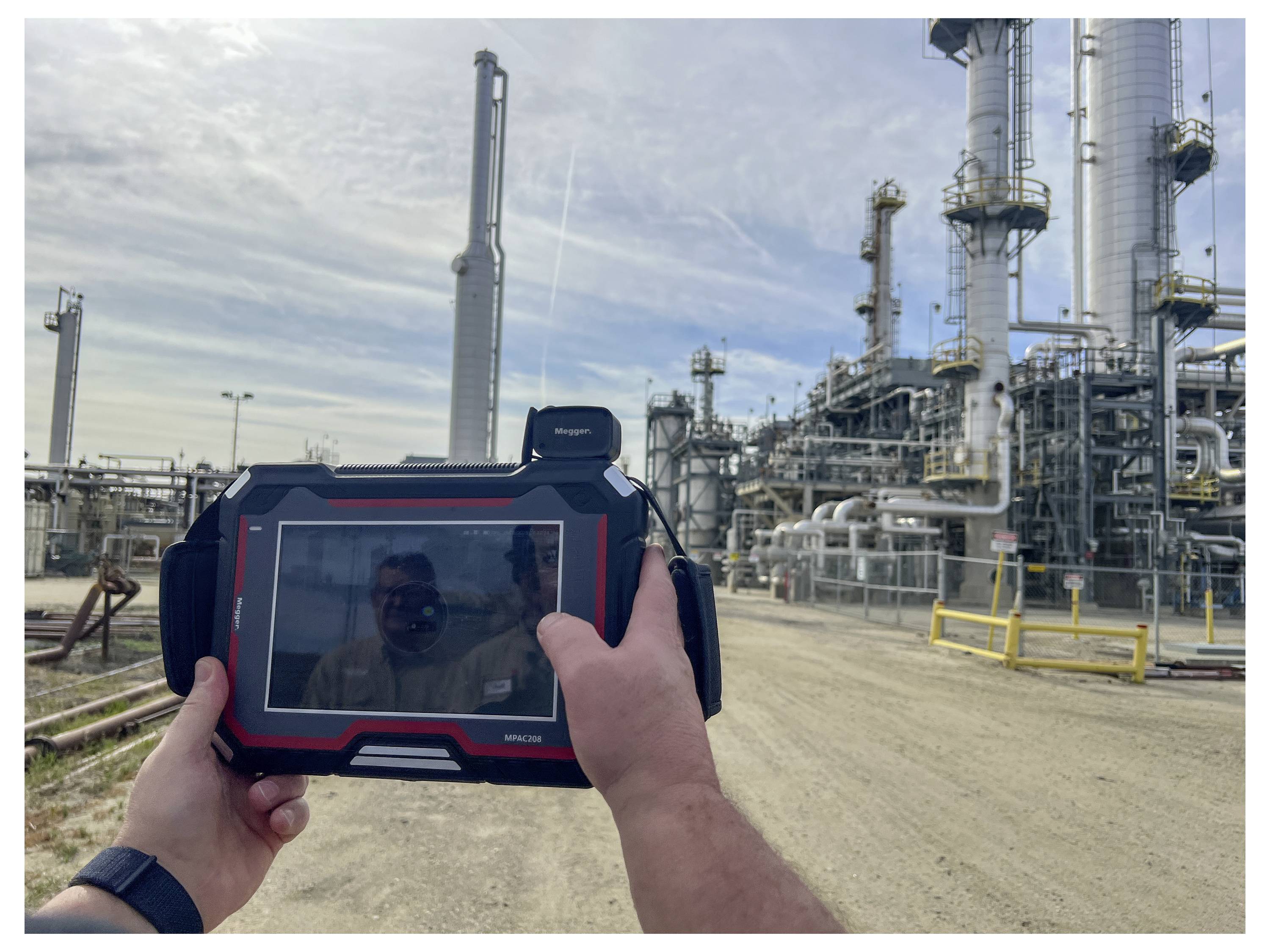 A person holds a tablet with a visible industrial plant in the background, capturing the facility's image and possibly monitoring data.