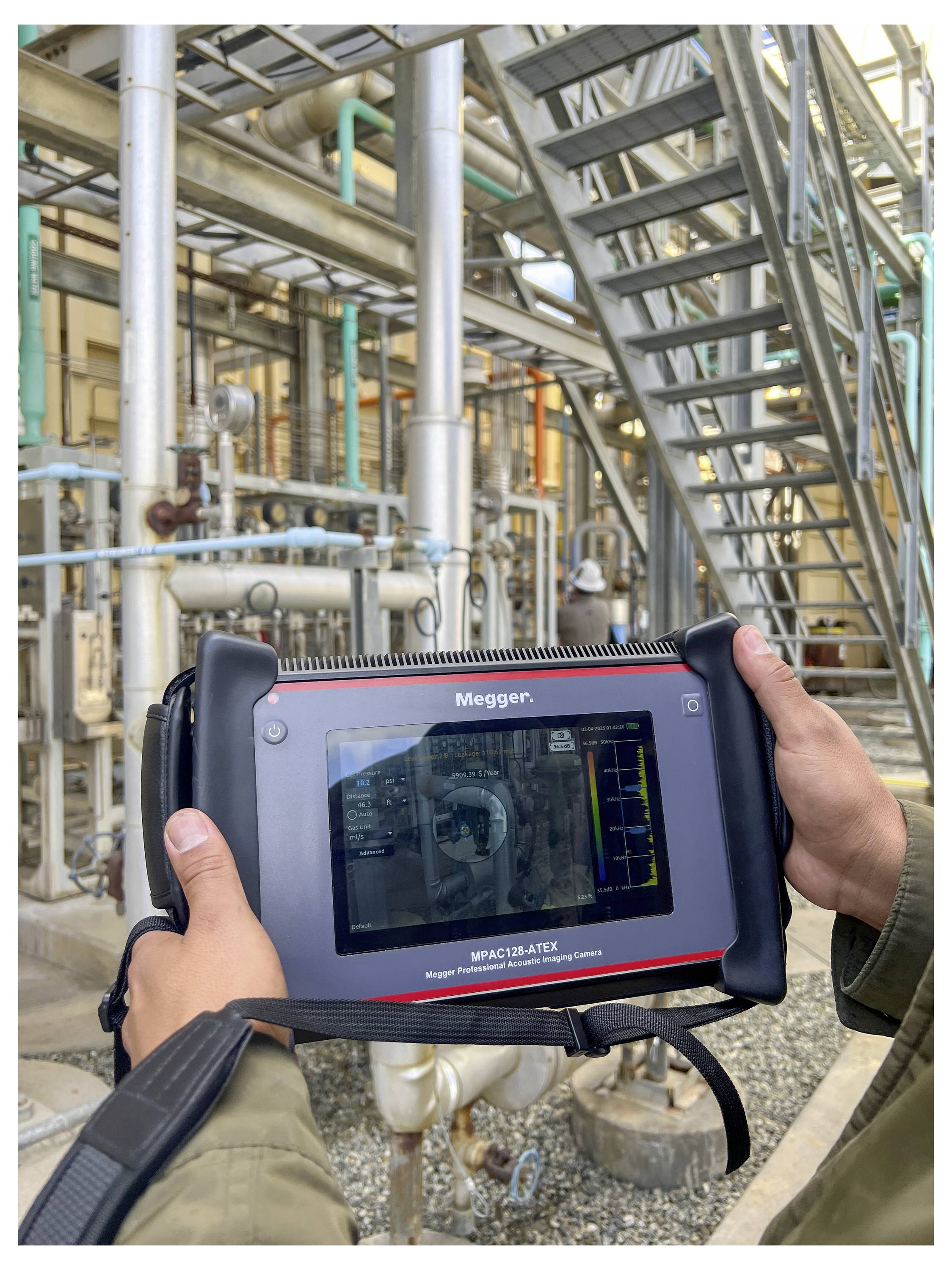 A person holds a diagnostic device with a screen displaying technical data, used for assessing industrial equipment, amidst complex piping.