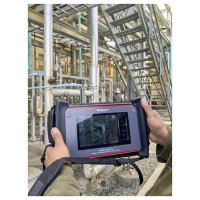 A person holds a diagnostic device with a screen displaying technical data, used for assessing industrial equipment, amidst complex piping.