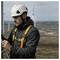 A worker wearing a helmet and safety gear stands on a wind farm, inspecting equipment with wind turbines visible in the background.