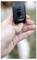 A hand holding a black GPS tracking device outdoors, possibly for a pet or person, with a green status light indicating it's active.