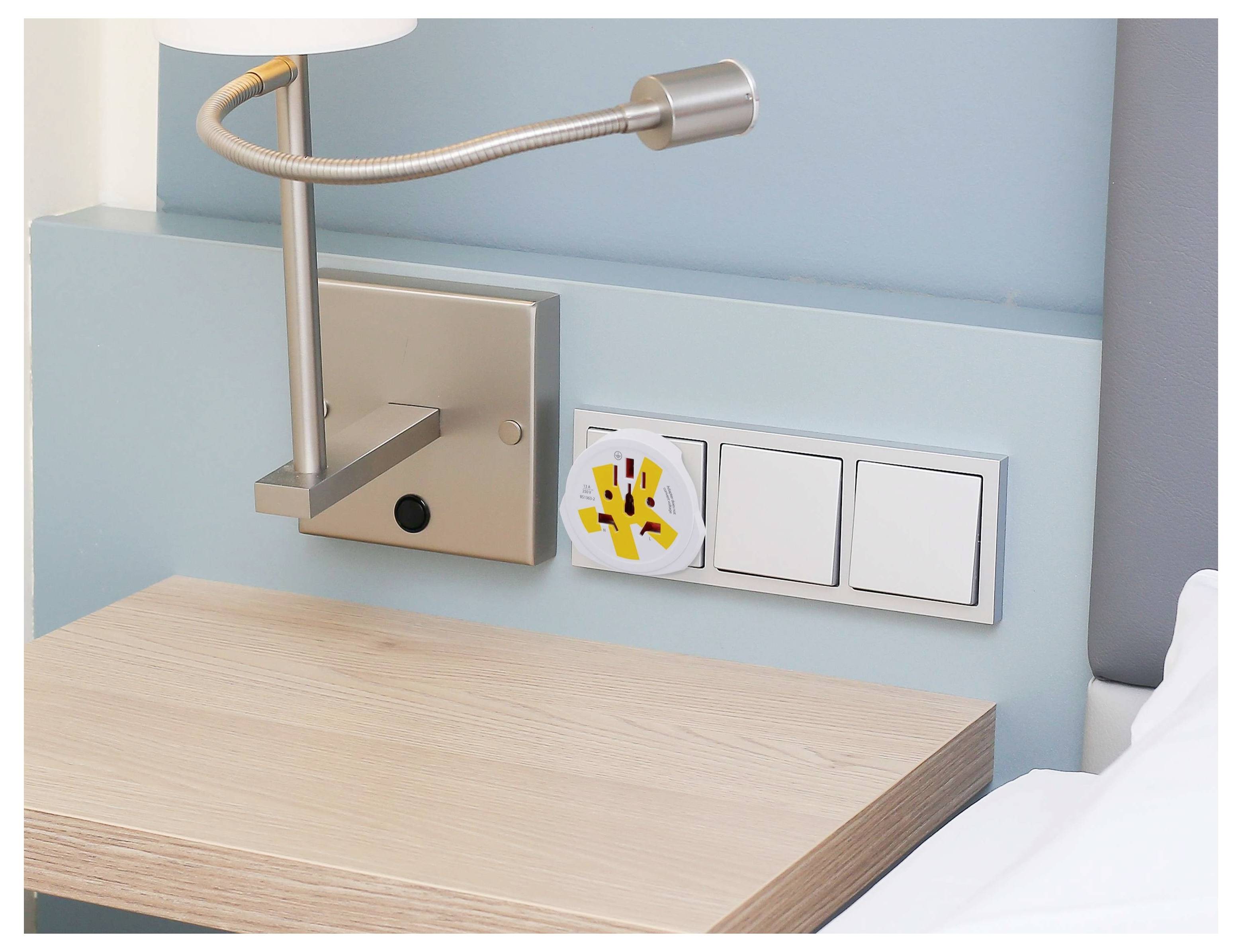 A bedside lamp on a light blue wall is next to a wall socket featuring a safety cover with a child hand warning symbol.