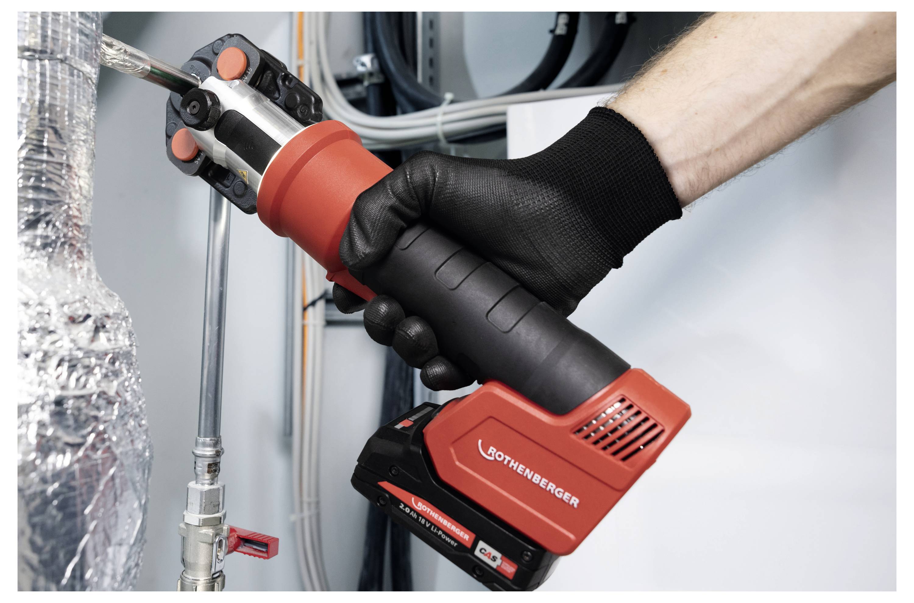 A gloved hand holds a red and black pipe press tool, pressing a plumbing pipe joint in an industrial setting.