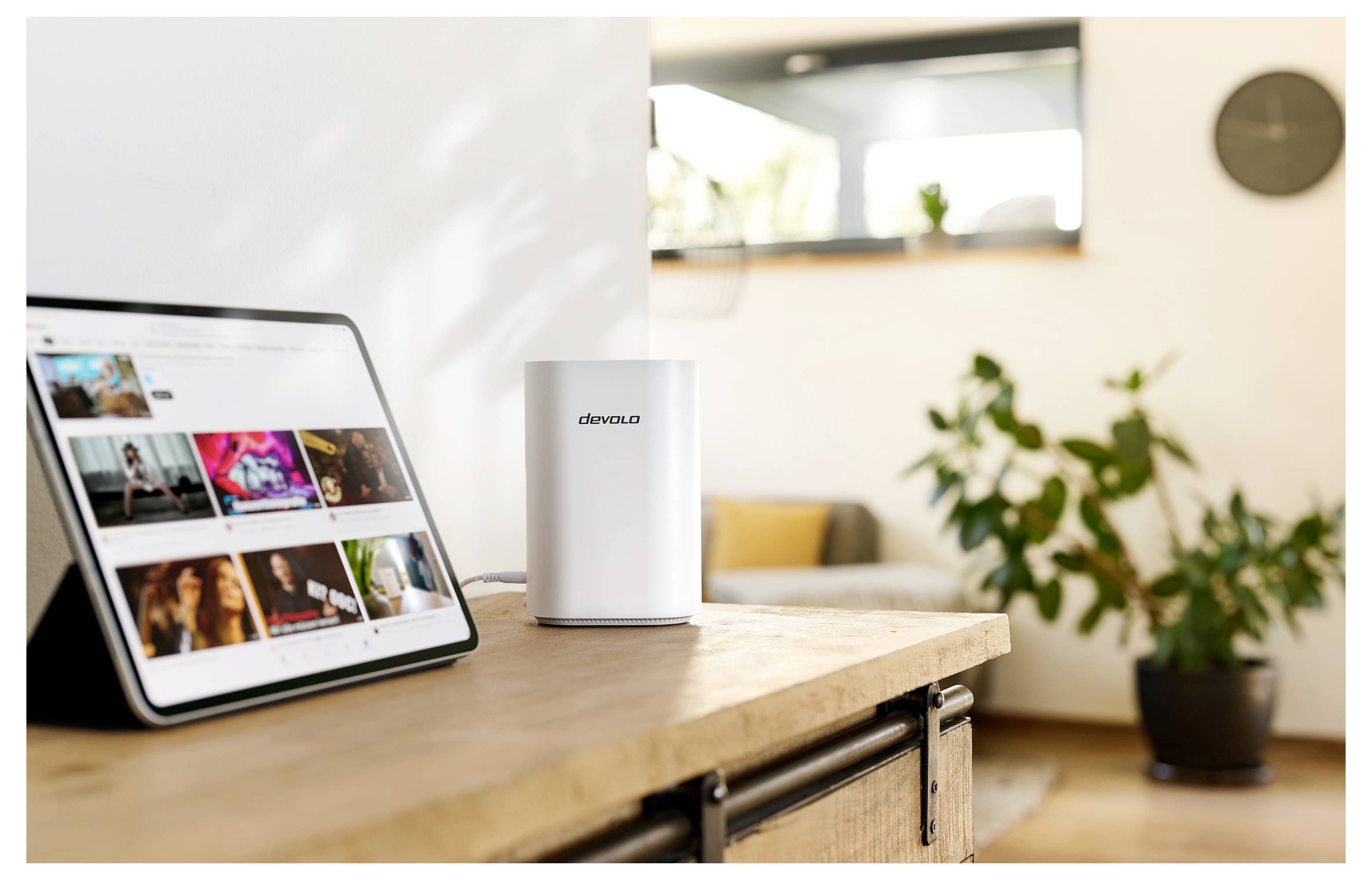 A tablet displaying a video library on a wooden table beside a white router, with a potted plant and couch in the background.
