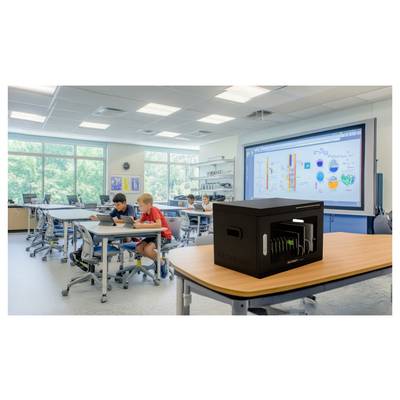 3D printer on a classroom table in a modern science lab, with students working at desks and a large data display in the background.
