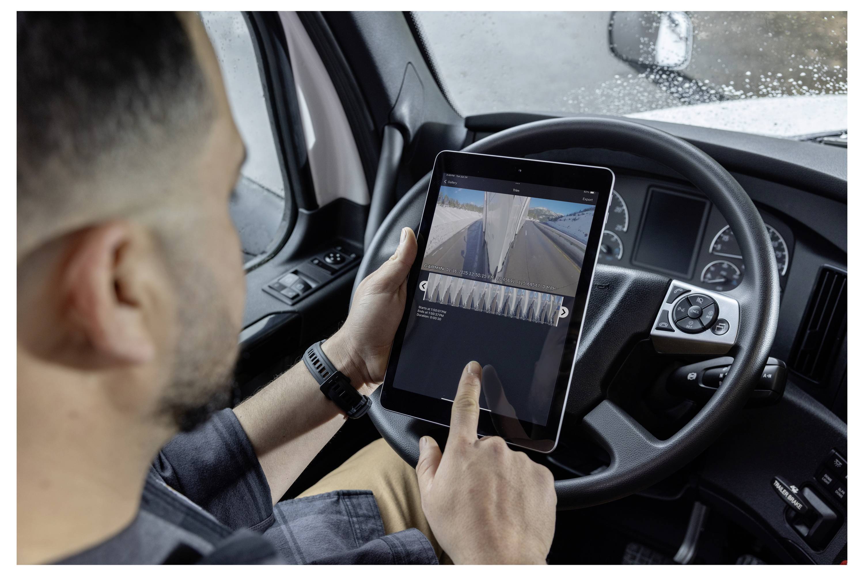 A person inside a truck uses a tablet to view a rear-view camera feed while parked. The truck's steering wheel and dashboard are visible.