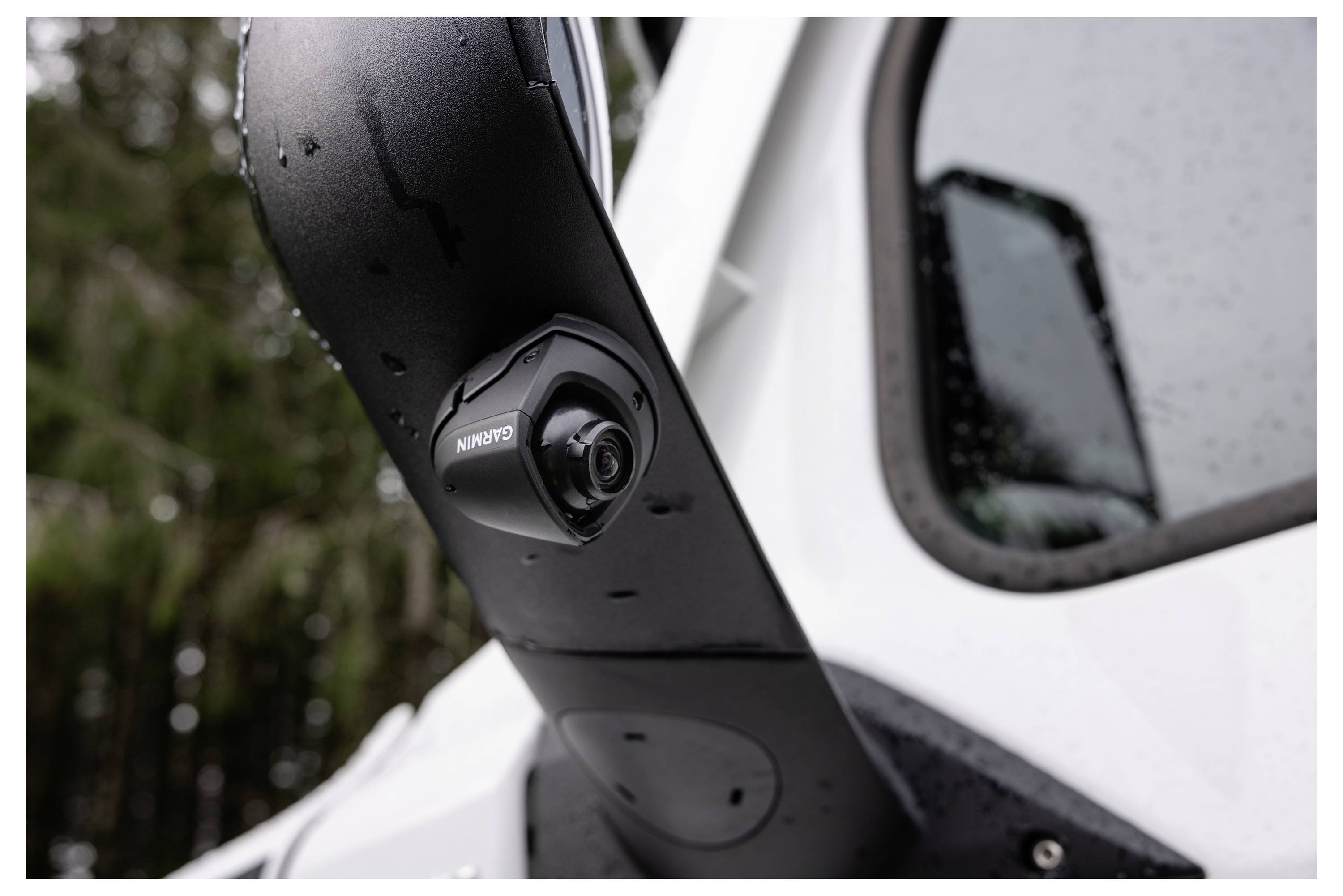 A side-view mirror with an integrated camera on a vehicle, featuring the branding 'Garmin', with trees reflected on its wet surface.
