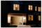 A house at night with lit windows and an open garage. A robot vacuum is visible inside the garage.