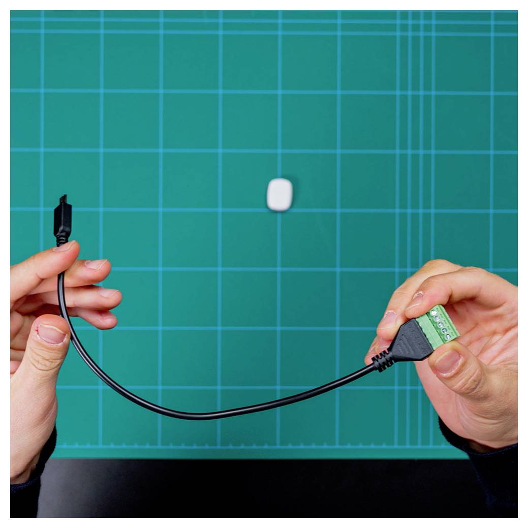 Two hands hold a black USB cable above a green grid-lined cutting mat. A small white rectangular object is on the mat.