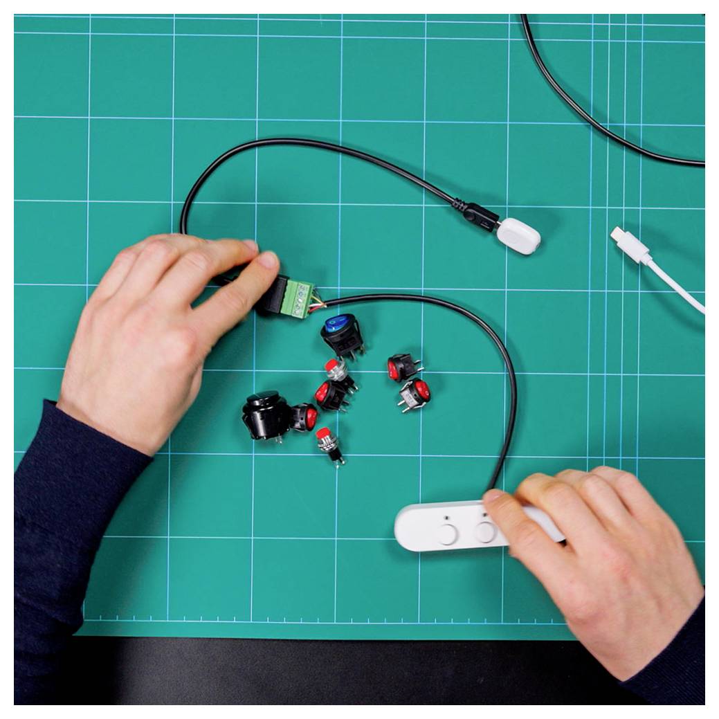 Two hands assembling electronic components on a cutting mat, including wires, connectors, and a control module, indicating a DIY electronics project.