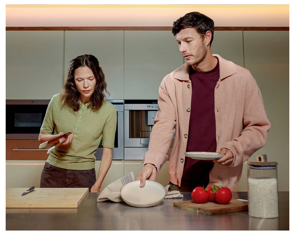A man and woman stand in a kitchen. The man arranges dishes beside tomatoes and cereal, while the woman looks at a smartphone.