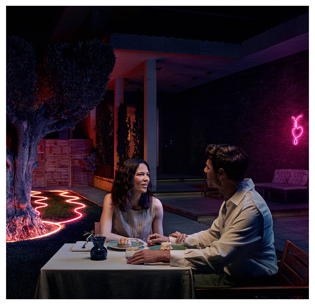 A couple sits at an outdoor restaurant table under evening lights, conversing. A neon light decorates the background.
