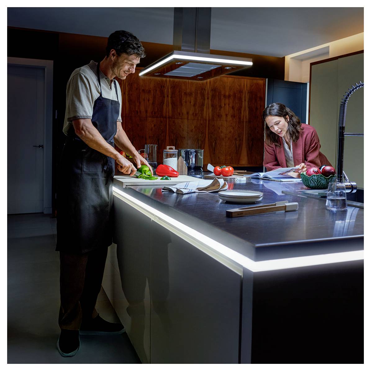 'A man chopping vegetables while a woman reads a cookbook in a modern kitchen.'