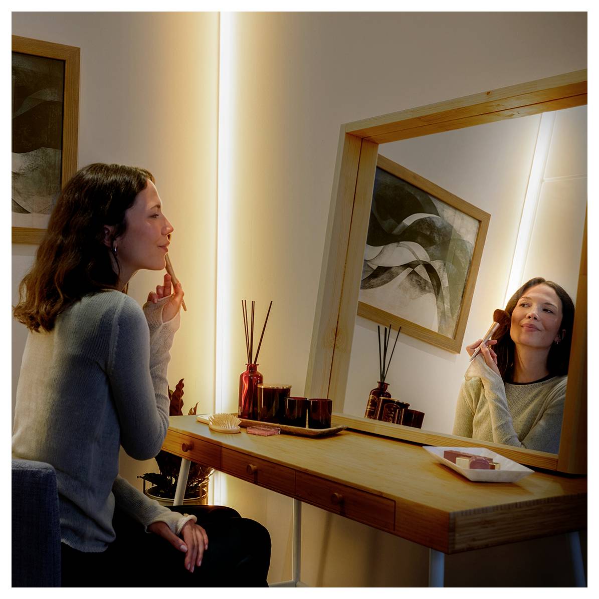 A woman seated at a vanity applies makeup while looking in a mirror, with abstract art on the wall and makeup items on the table.