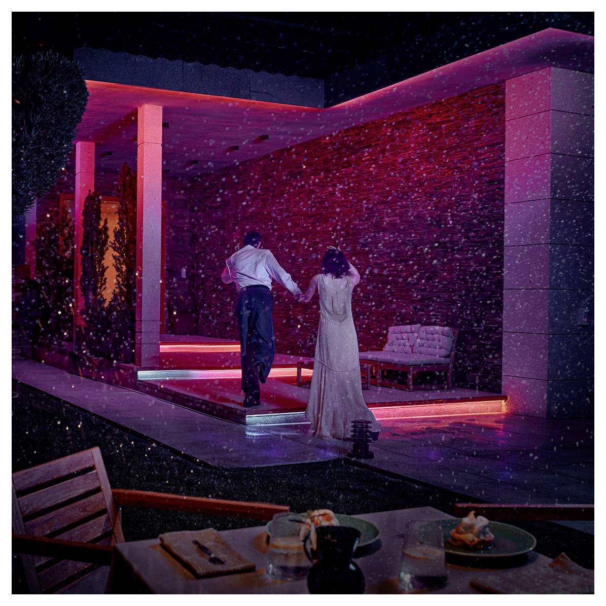 A couple, seen from behind, walks hand in hand in front of a building with pink neon lights and snow falling at night.