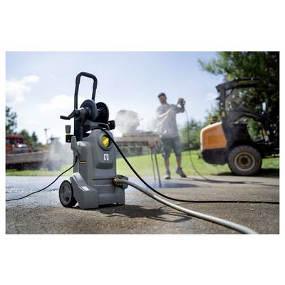 A person uses a pressure washer on a driveway, with a hose attached to it. A vehicle and trees are visible in the background.