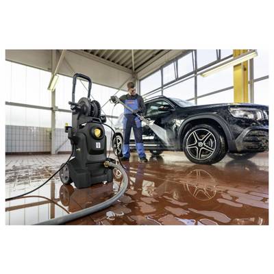 Person in blue overalls uses a pressure washer to clean a black car in a bright garage with tiled floor and large windows.
