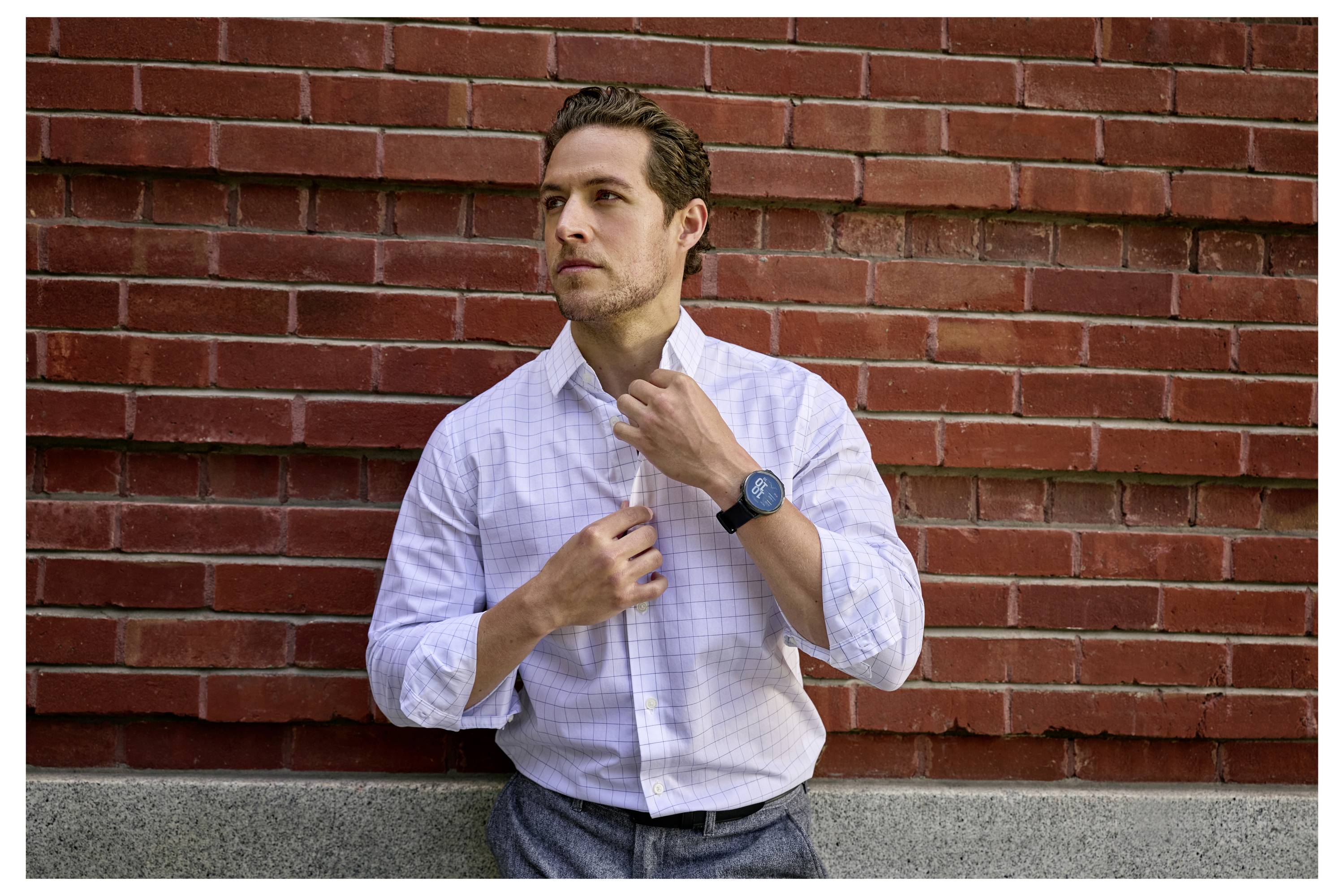 A person adjusts the collar of their white shirt while standing in front of a red brick wall, wearing a dark wristwatch.