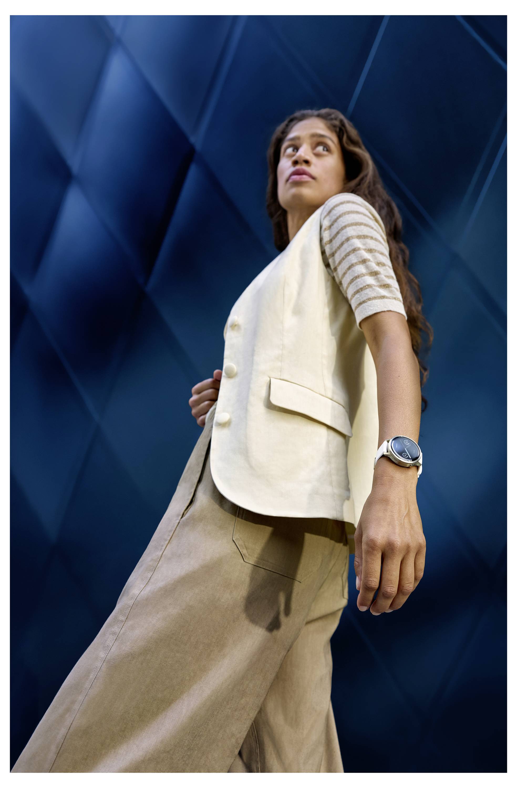 A person stands confidently gazing upward, wearing a white vest, striped shirt, and beige pants against a textured blue background.