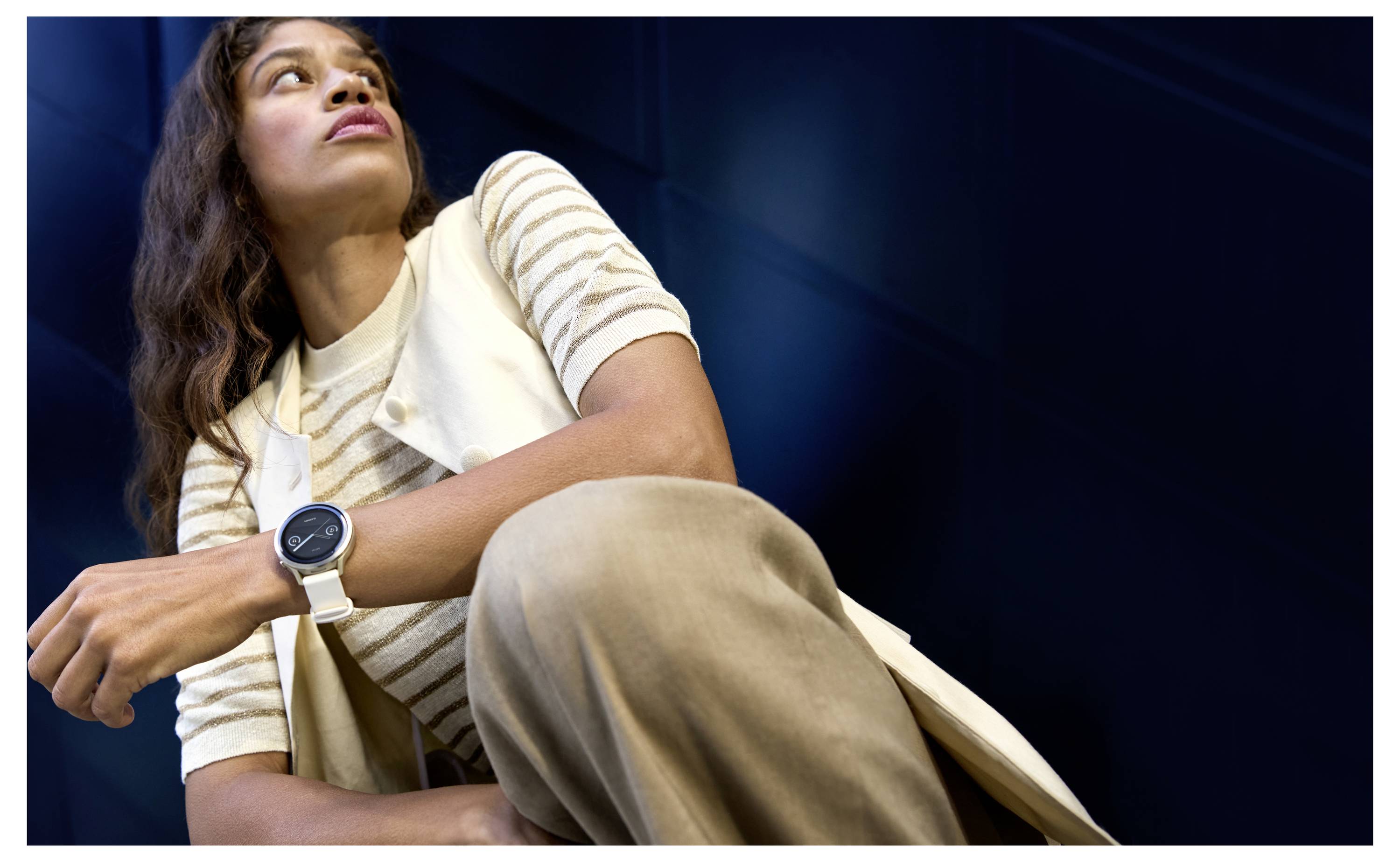 A person wearing a beige outfit and a white smartwatch, gazing upwards, against a dark background. The focus is on the watch.