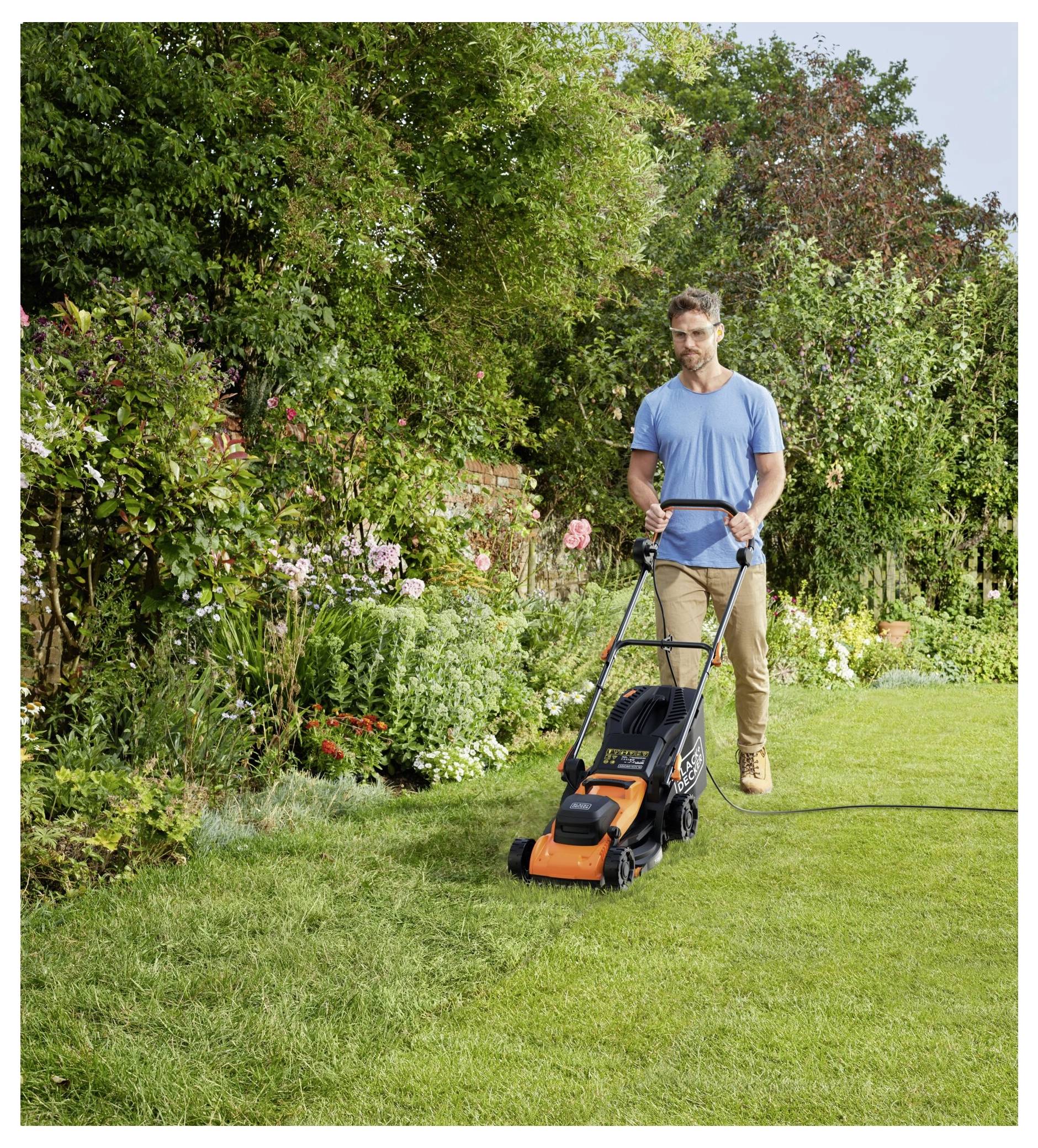 A person mowing a lush green lawn with an orange lawnmower in a garden surrounded by colorful plants and trees.