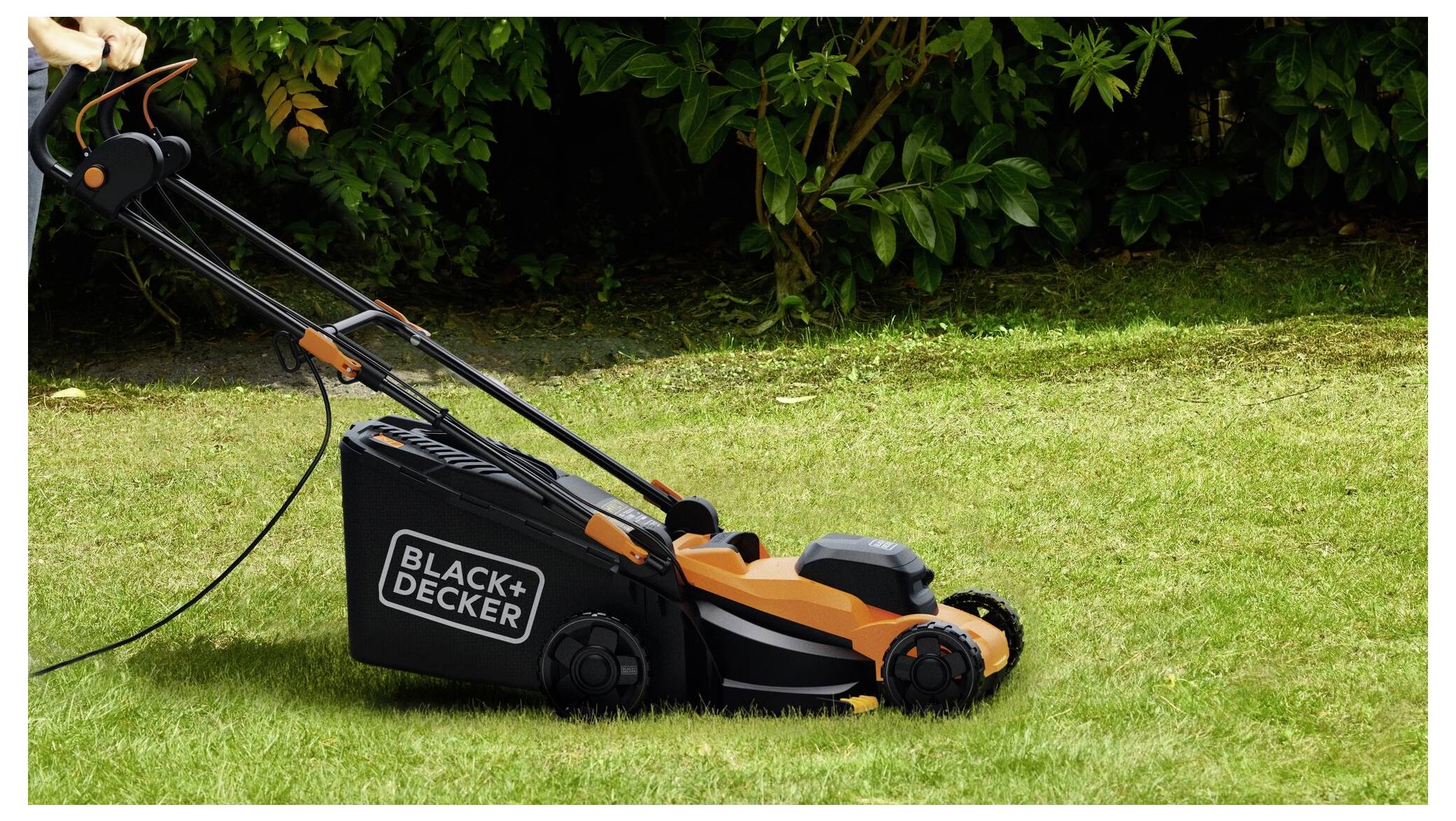 A person is pushing a Black+Decker lawnmower across a green lawn, with bushes in the background. The lawnmower is black and orange.