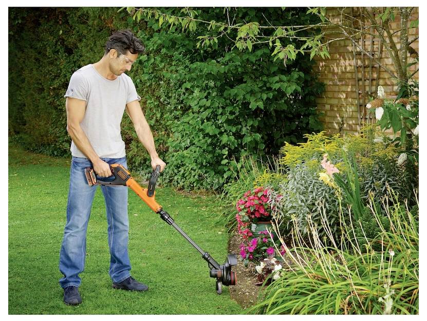 Person using an electric trimmer on garden edges, surrounded by colorful flowers and greenery, indicating outdoor landscaping work.