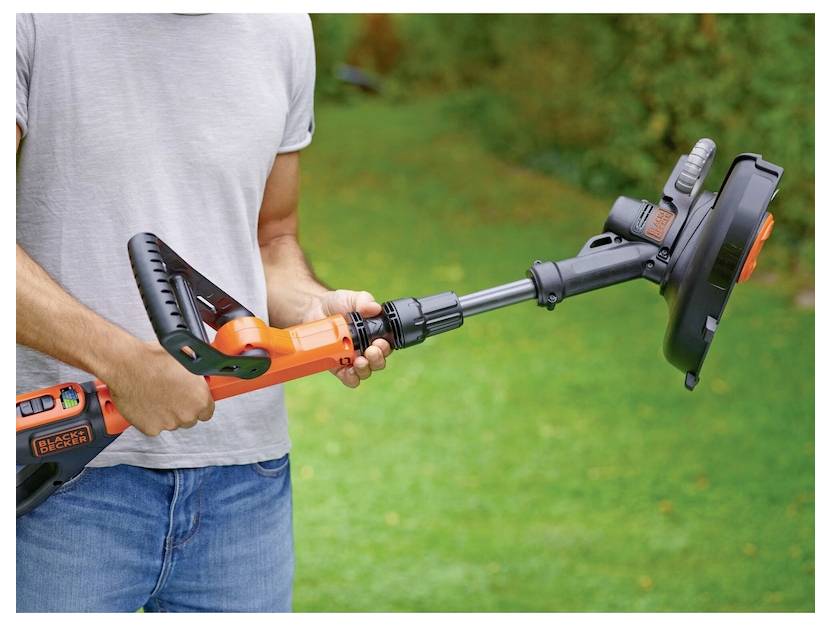 A person adjusts the length of an orange and black string trimmer in a grassy backyard, preparing it for use.