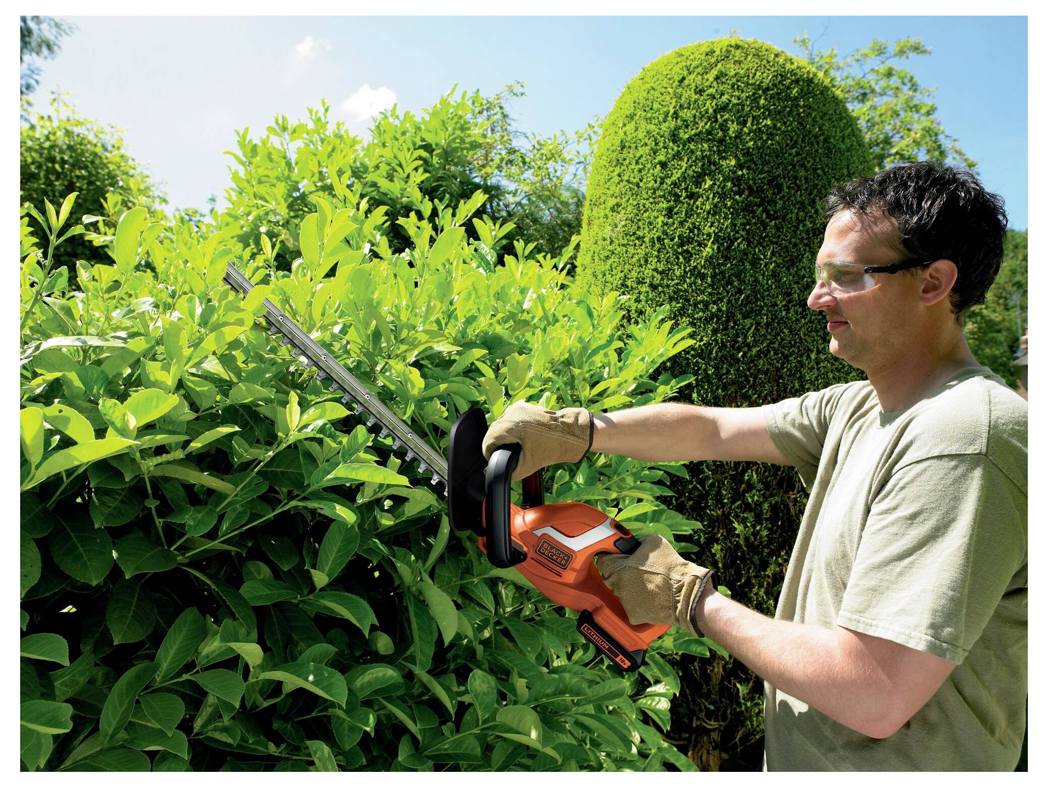 A person trims a hedge with an orange hedge trimmer, wearing gloves and goggles, outdoors on a sunny day with lush greenery.