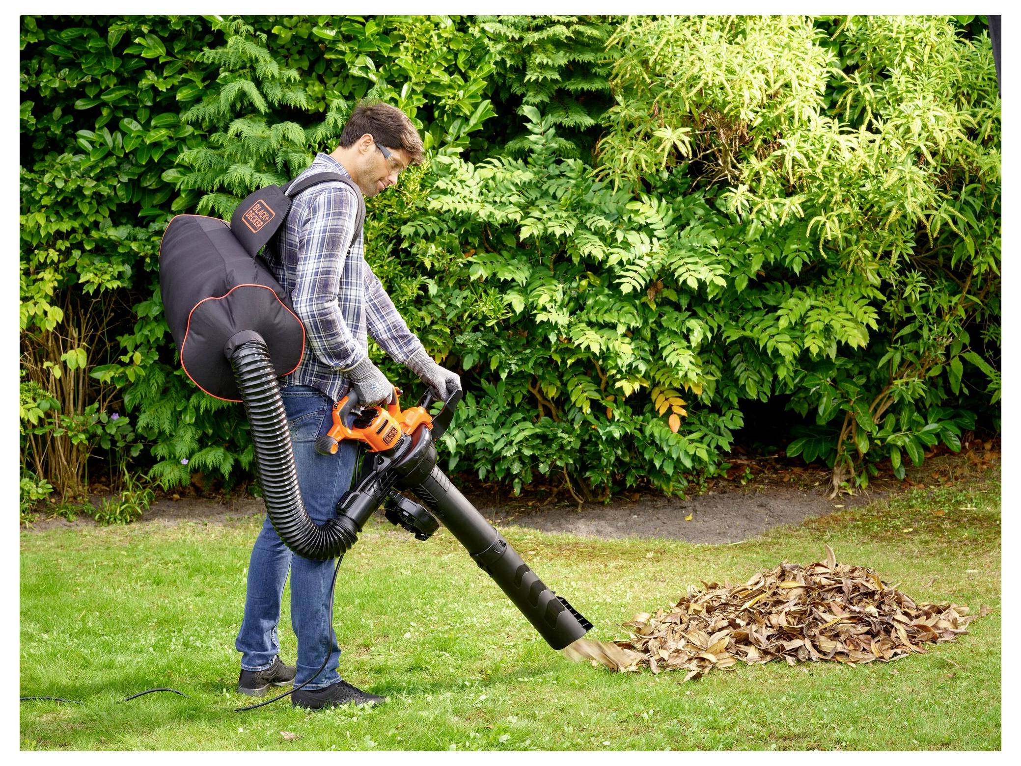 A person uses a leaf blower to collect fallen leaves into a pile on a grassy lawn, surrounded by lush green shrubs.