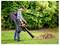 A person uses a leaf blower to collect fallen leaves into a pile on a grassy lawn, surrounded by lush green shrubs.