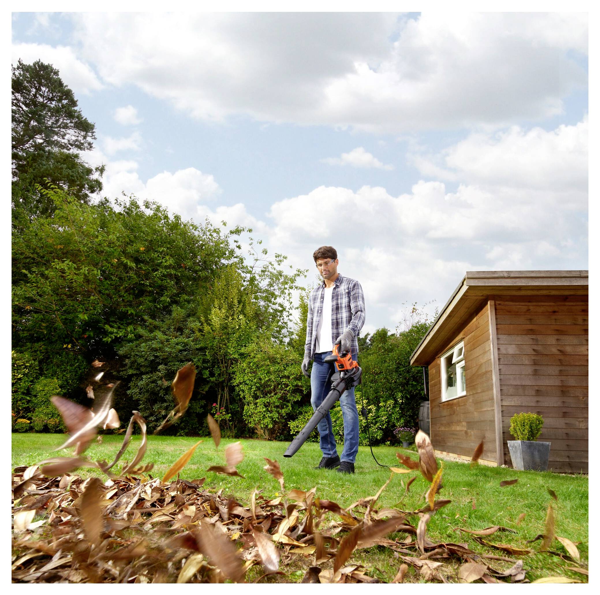 A man uses a leaf blower on a grassy yard, surrounded by tall trees and a small wooden shed, under a partly cloudy sky.
