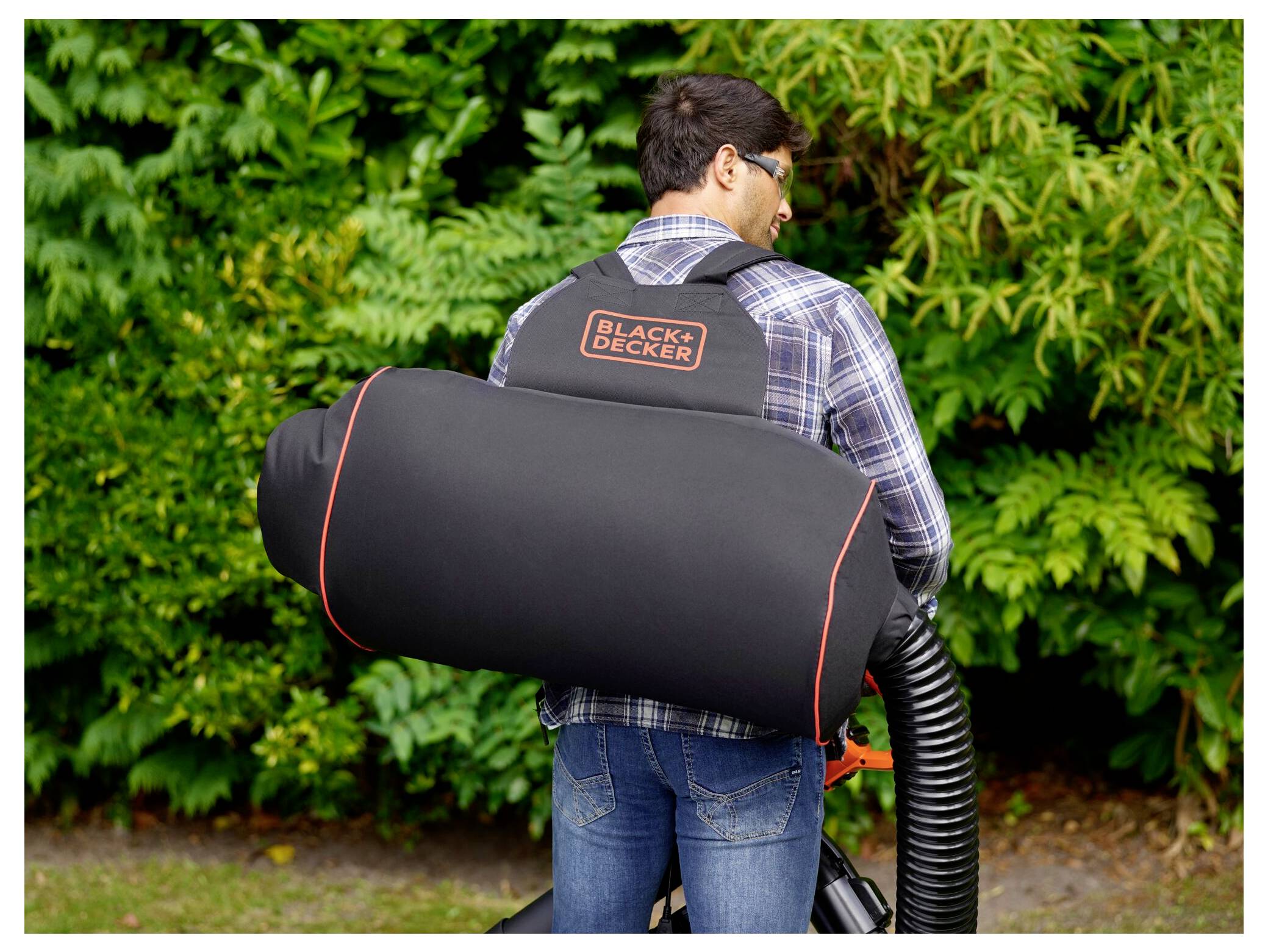 A person wearing a backpack leaf blower from Black & Decker stands in front of green foliage, preparing for garden maintenance.
