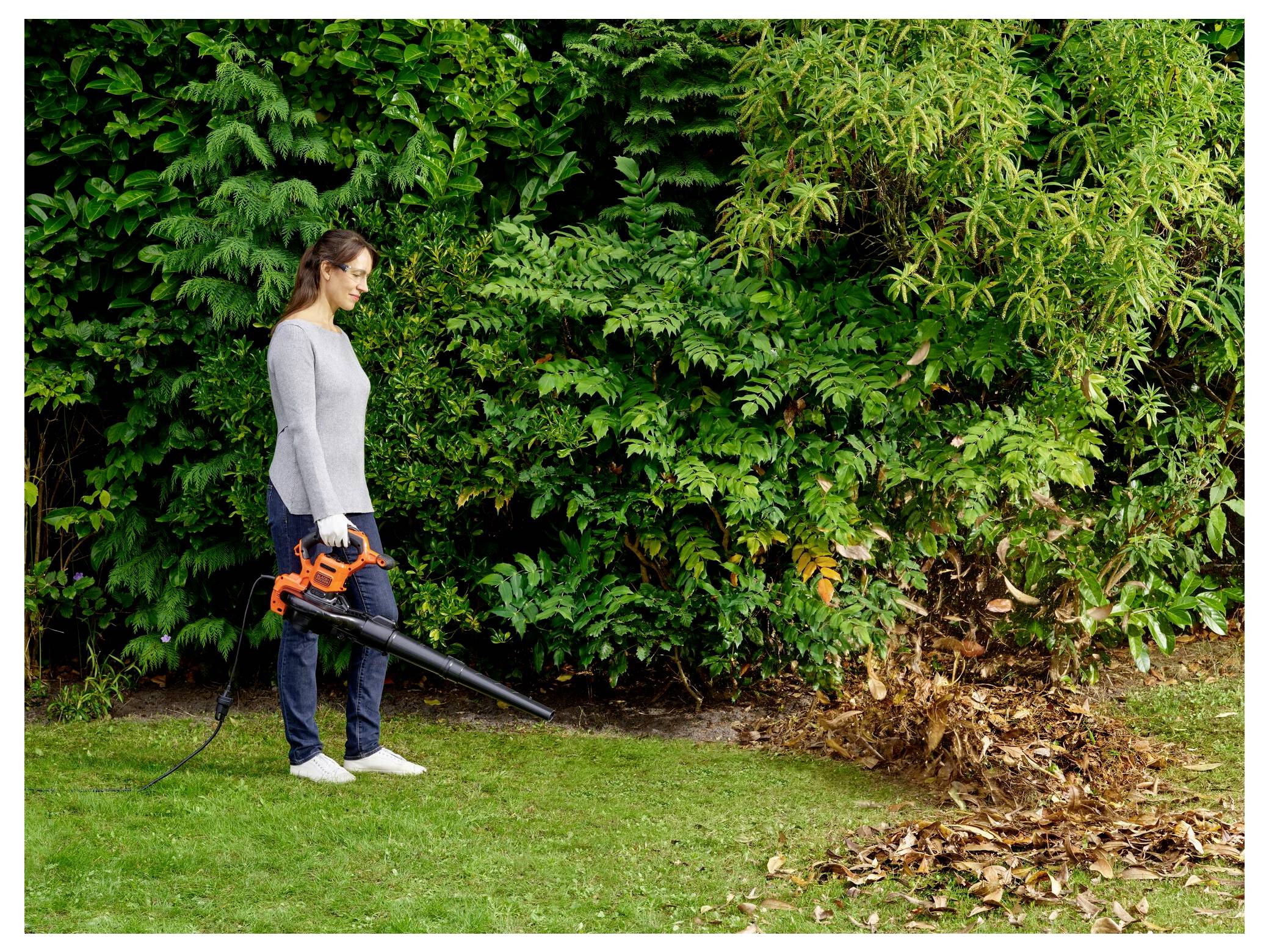 A person uses an electric leaf blower to clear a pile of leaves on a grassy area, with lush green bushes in the background.