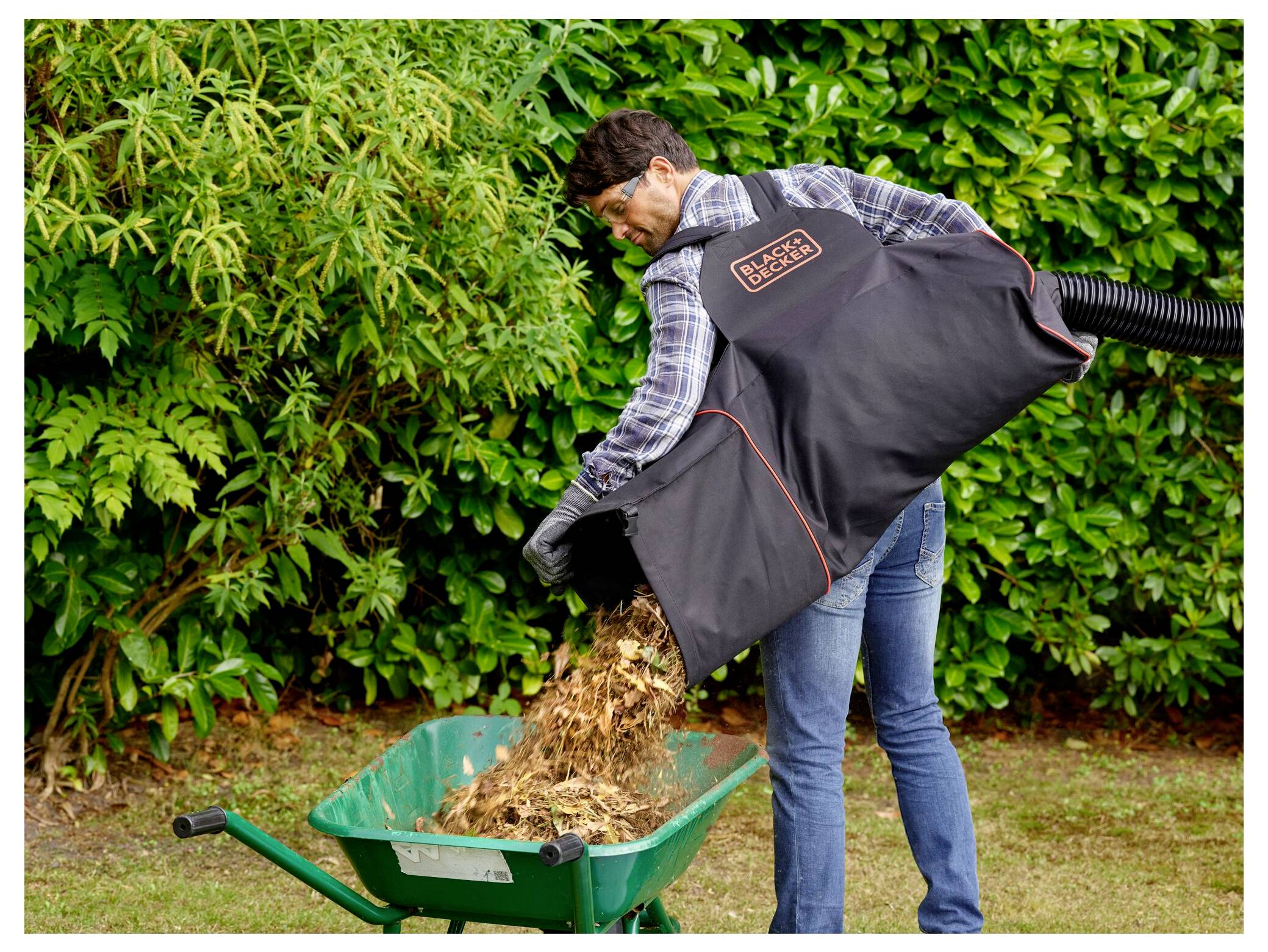 A person uses a leaf blower vacuum with a collection bag to dump shredded leaves into a green wheelbarrow, standing in a garden.