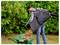 A person uses a leaf blower vacuum with a collection bag to dump shredded leaves into a green wheelbarrow, standing in a garden.