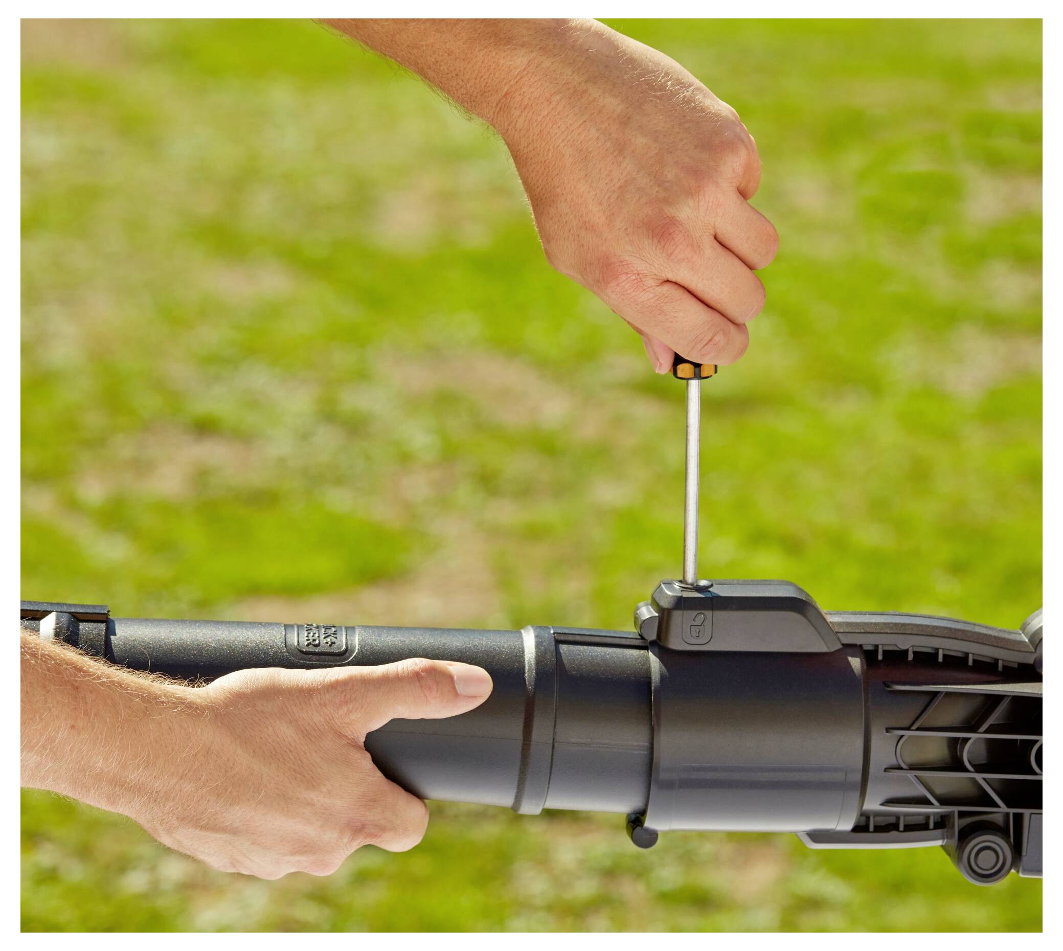 A person uses a screwdriver to assemble or adjust a garden tool on a sunny day in a grassy area.