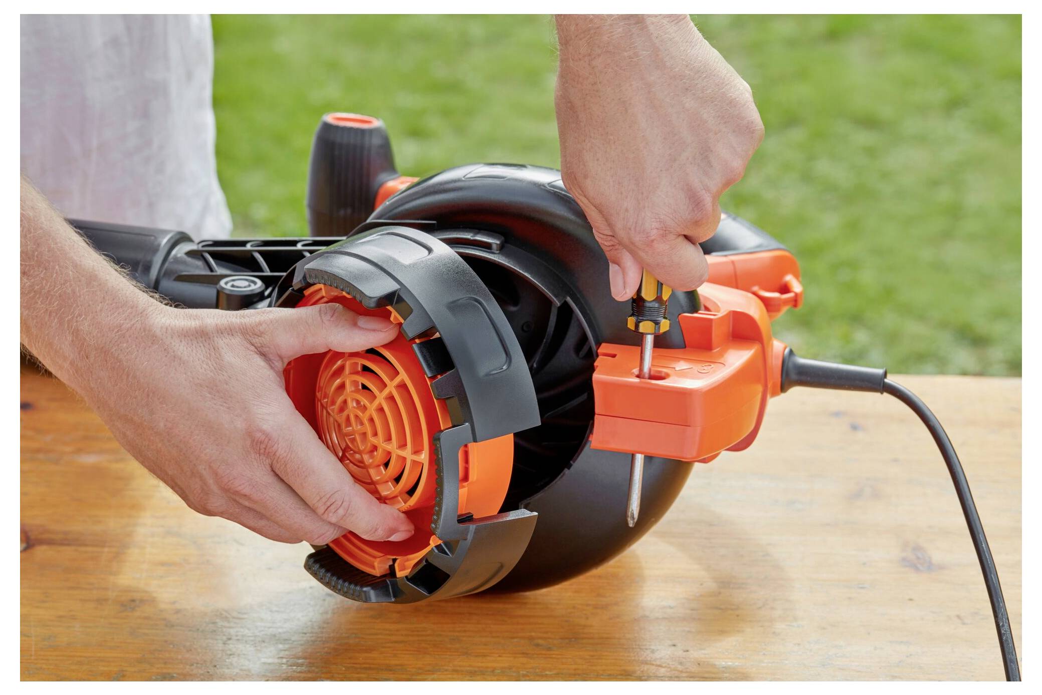 Person assembling an orange and black power tool on a wooden surface outdoors, focusing on tightening a component with a screwdriver.