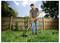 A person trims grass in a garden using an orange trimmer, standing in front of a wooden fence on a sunny day.