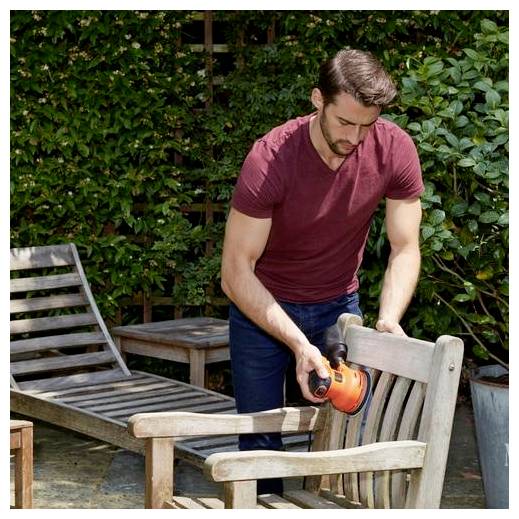 A person using a power sander on a wooden chair outdoors. Green bushes in the background, setting indicates yard work or DIY project.