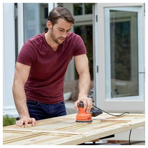 A person in a red shirt uses an orange electric sander on a wooden door placed on a table outdoors.