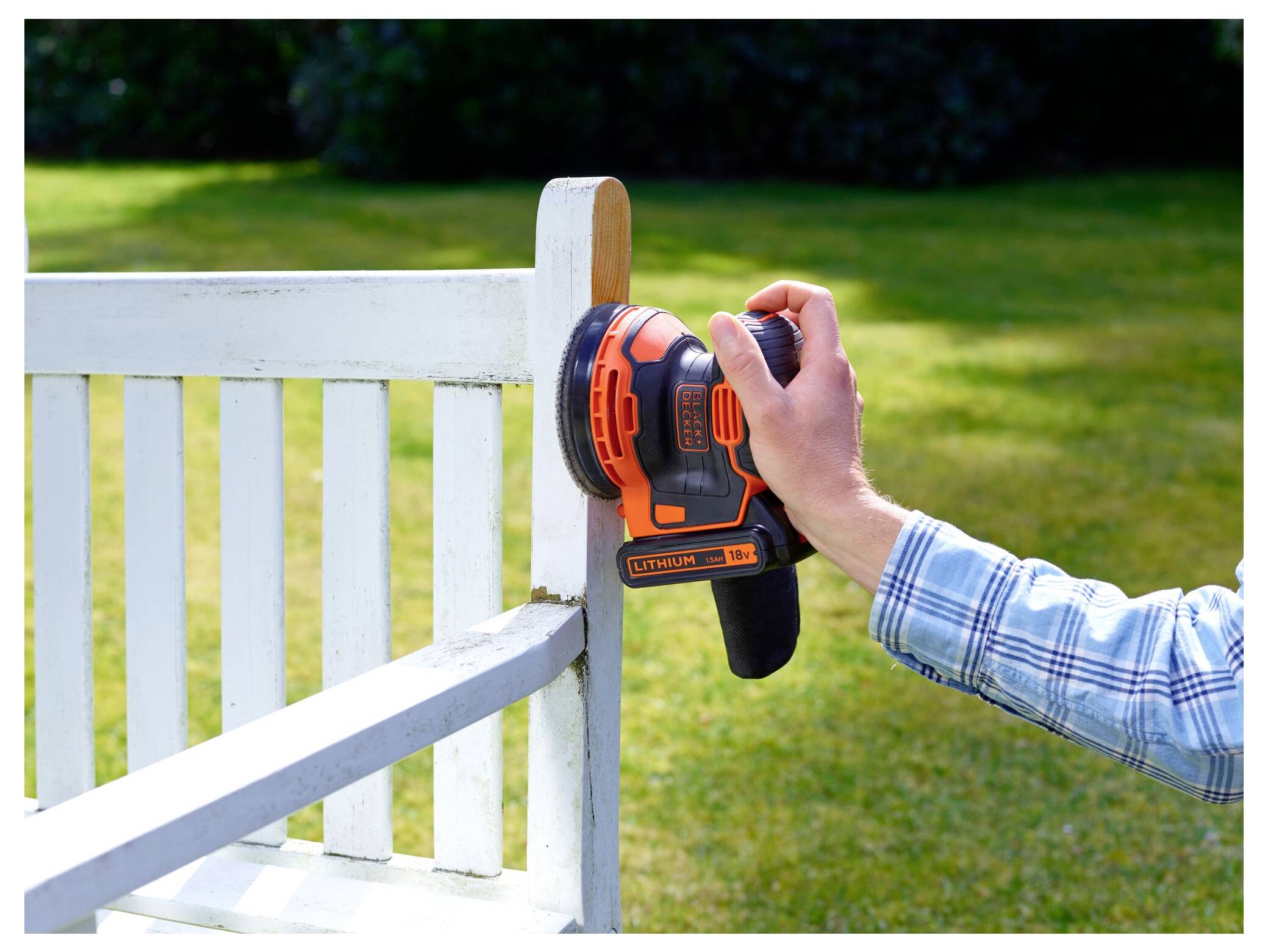 A person uses an electric sander on a wooden bench in a sunny garden, suggesting outdoor furniture maintenance or renovation.