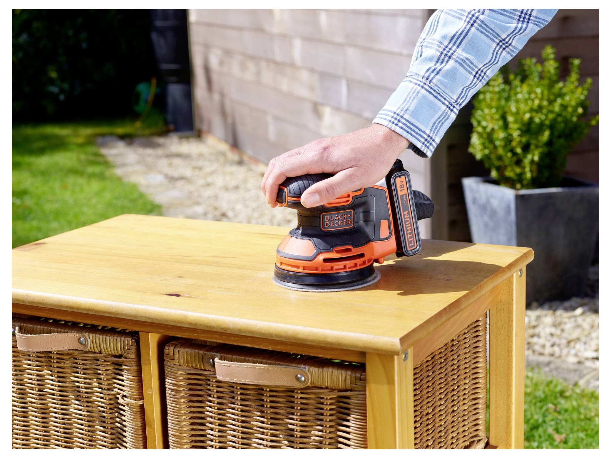 A person in a checkered shirt uses an orange and black sander to smooth a wooden cabinet on an outdoor patio.
