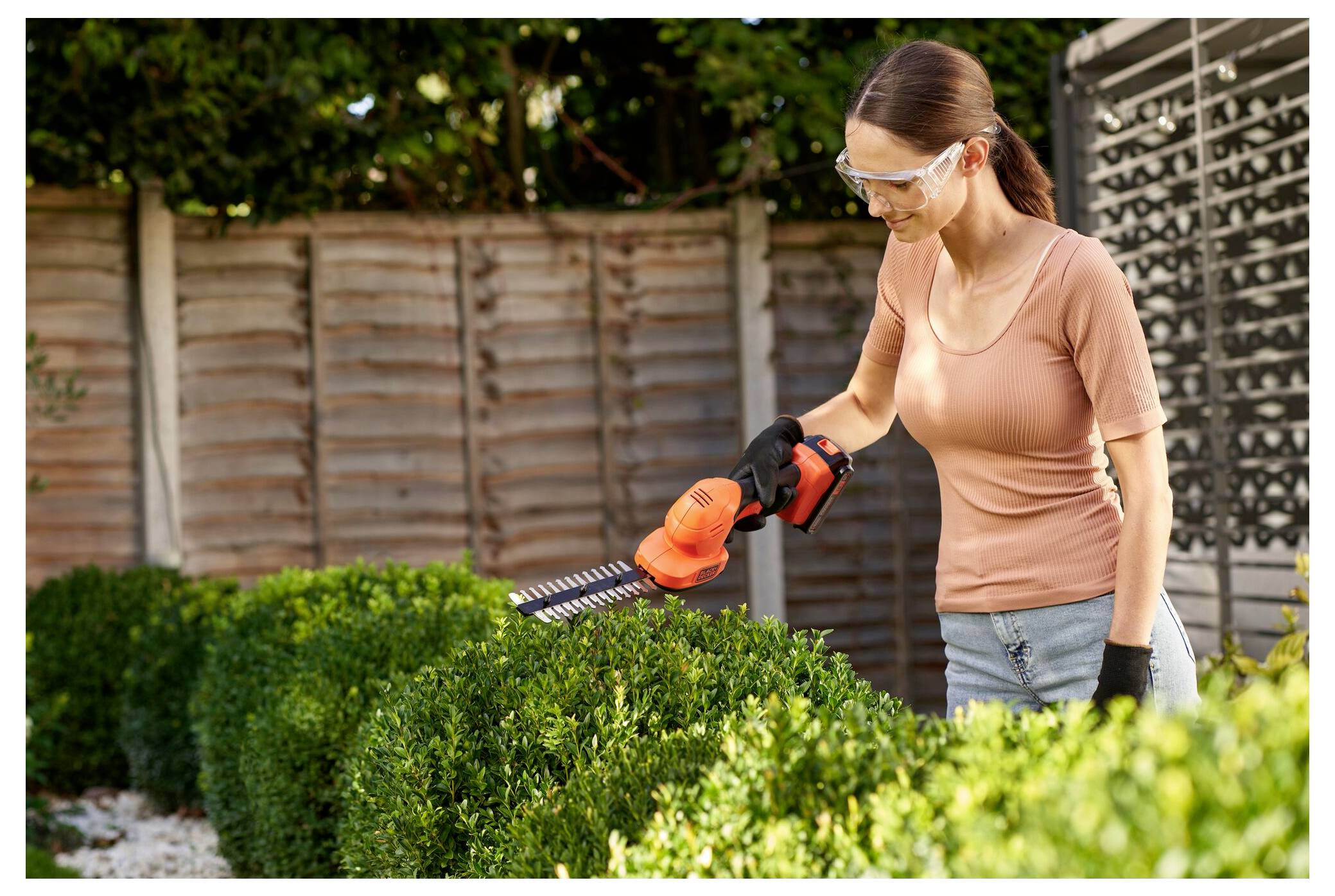 A person wearing goggles and gloves uses an electric hedge trimmer to shape a neatly trimmed shrub in a garden with a wooden fence.