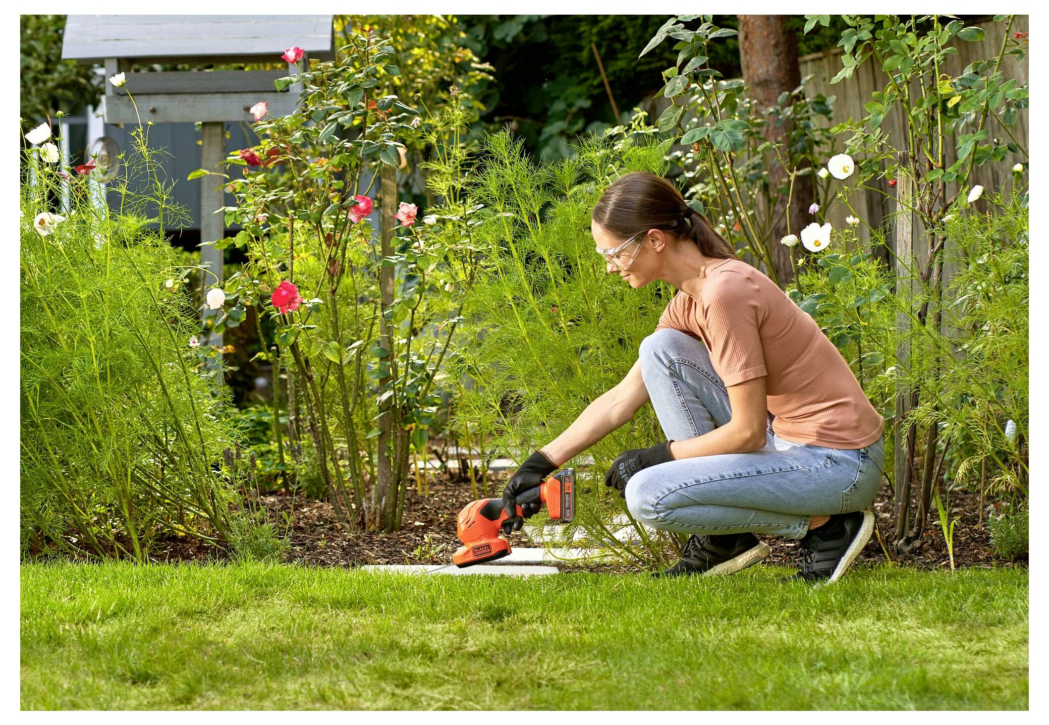 A person wearing safety glasses is kneeling in a garden, using an orange trimmer to cut plants, surrounded by flowers and greenery.