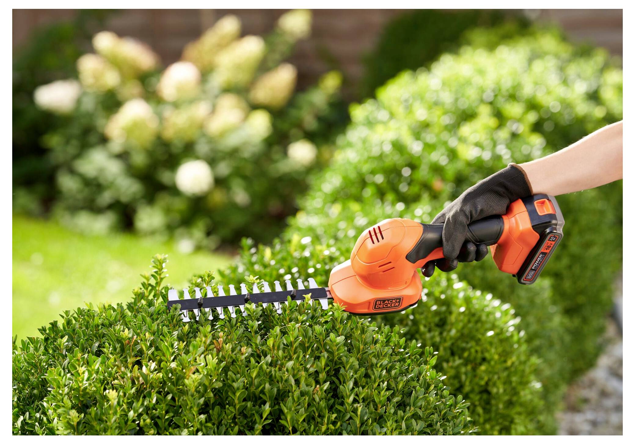 A person using an orange handheld electric hedge trimmer to shape a well-manicured bush in a garden with blurred flowers in the background.