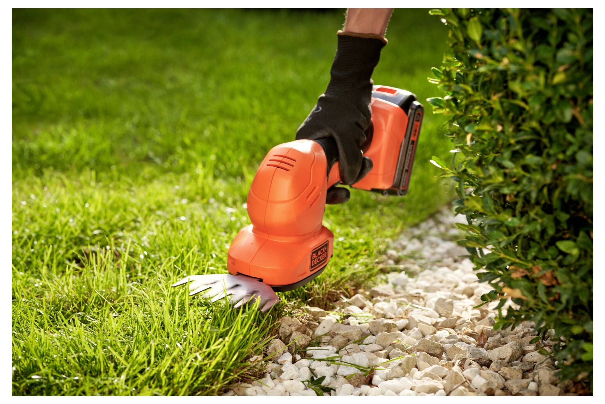 A person trims the edge of a lawn with an orange hand-held grass shear next to a hedge and gravel path.