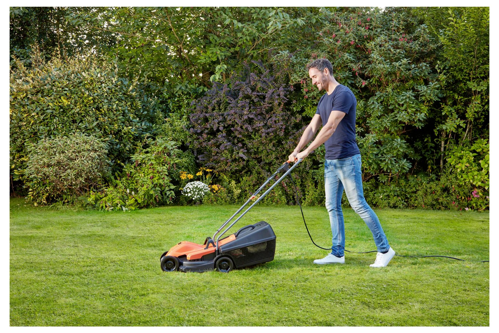 A person mowing a green lawn with an electric lawnmower in a garden, surrounded by bushes and trees under a clear sky.